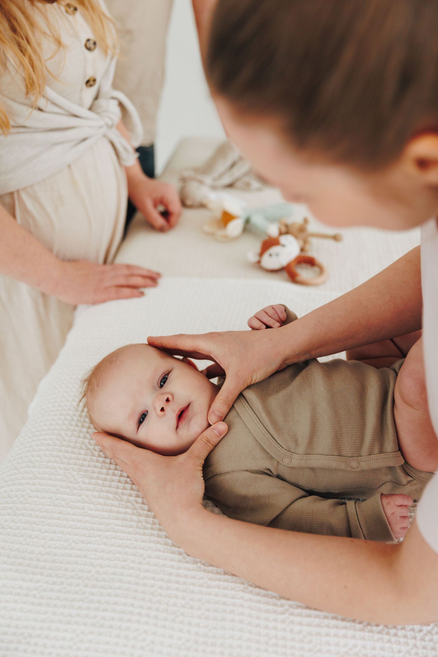 A healthcare professional gently holds a baby in an olive romper during a pediatric checkup with a pregnant woman nearby.