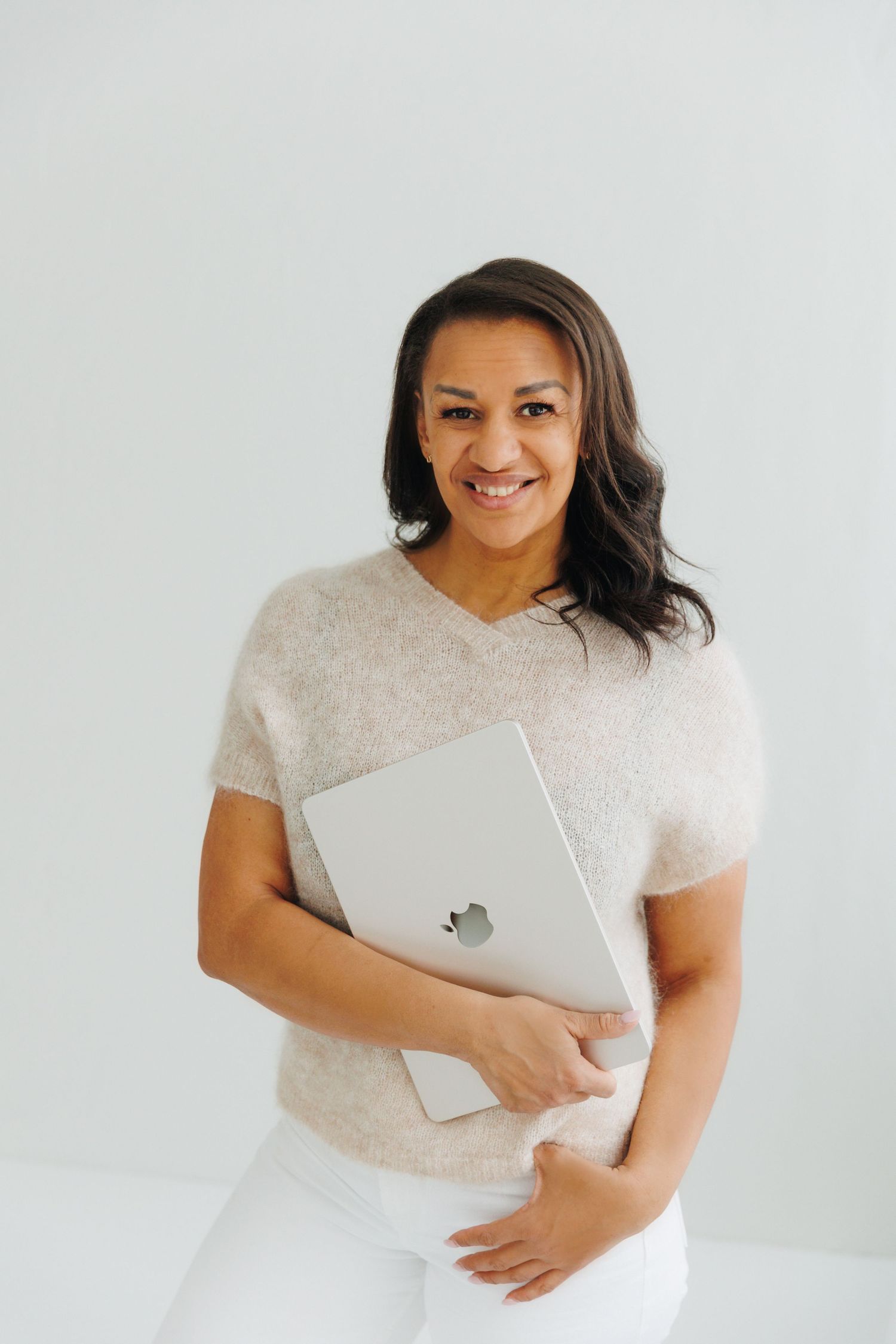 Smiling woman holding a silver Apple laptop, wearing a beige sweater and white pants against a light background.