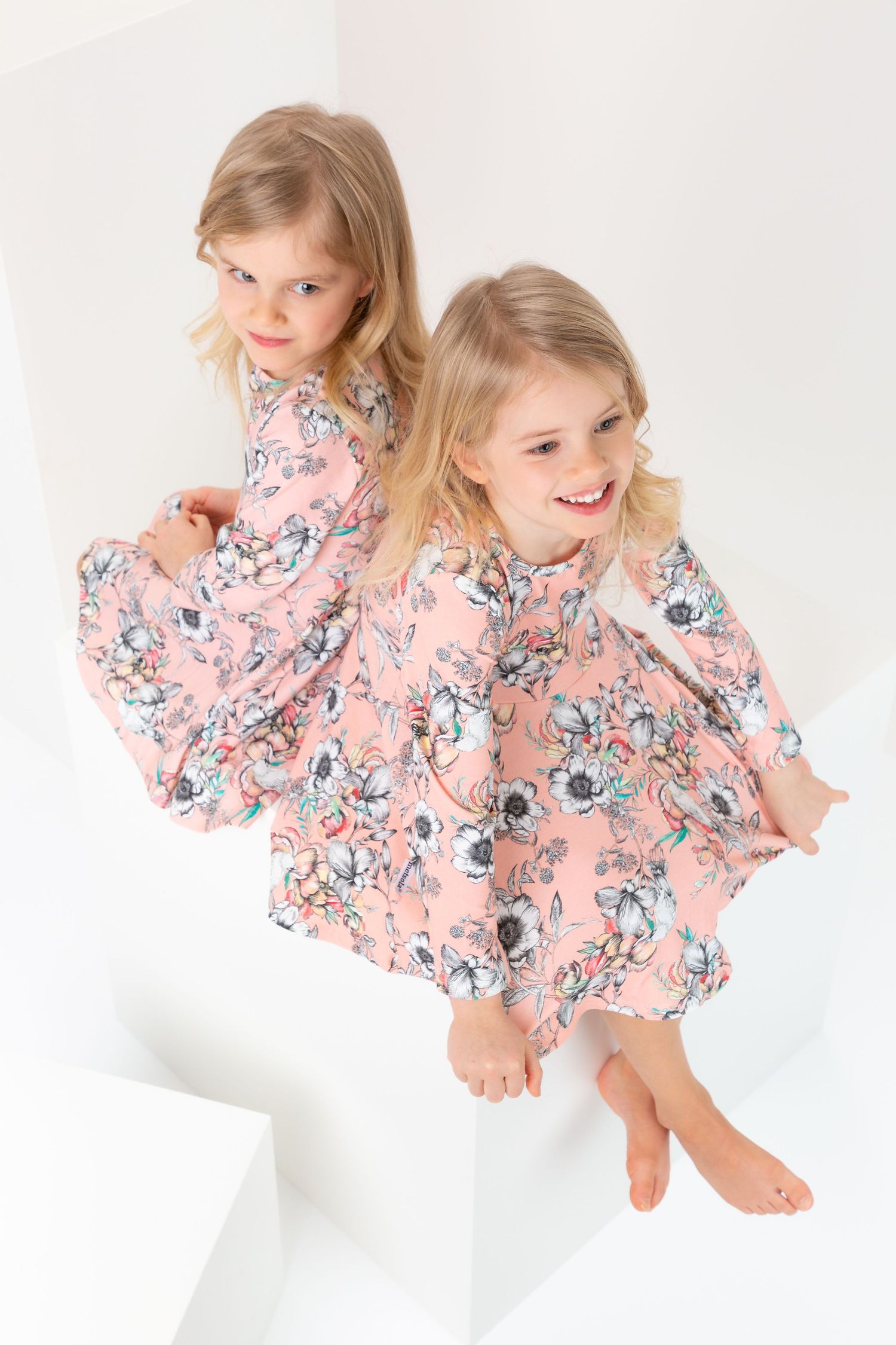 Two young girls in matching pink floral dresses sitting together on white steps, smiling and barefoot.