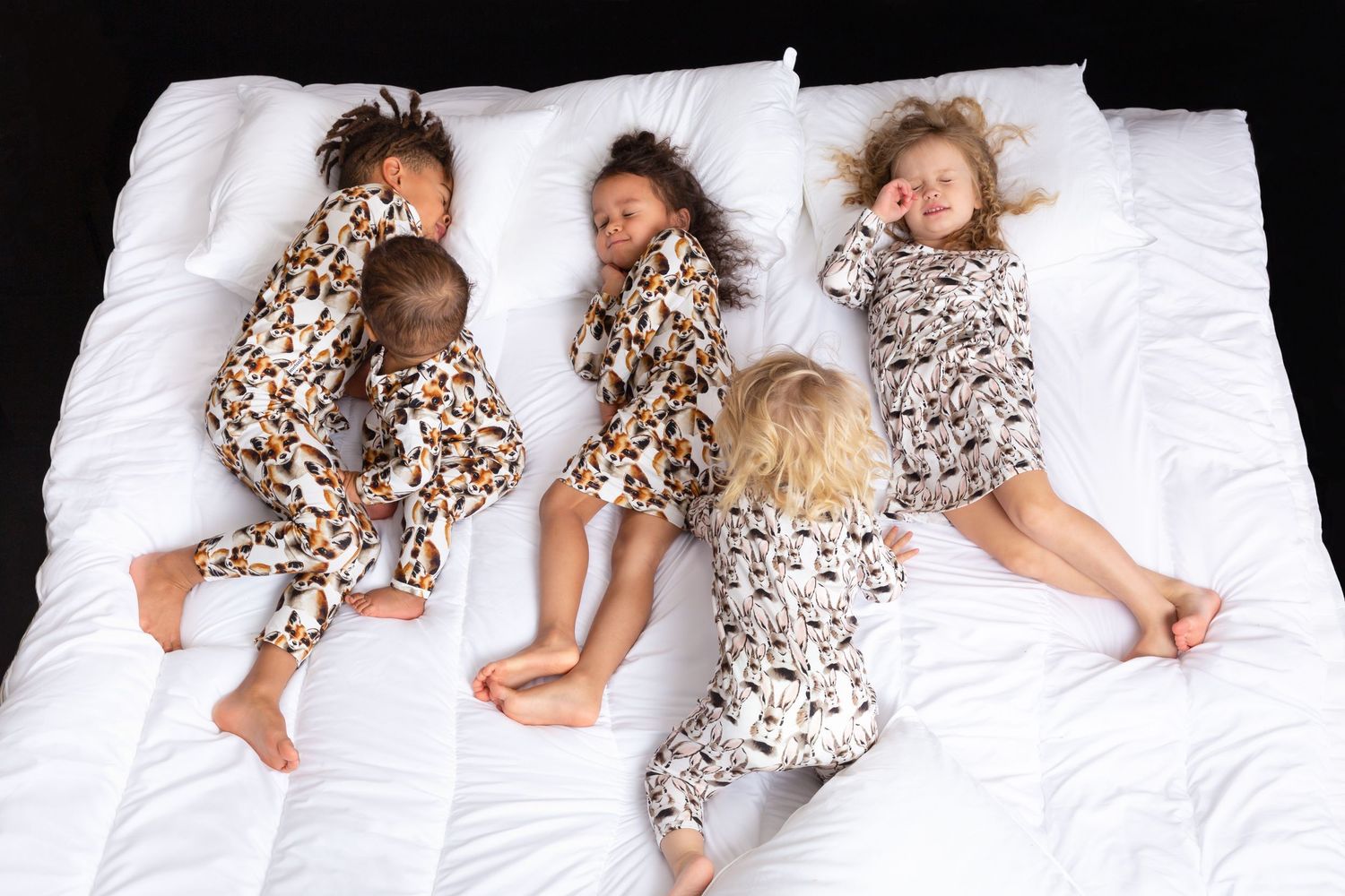 Four children in matching printed pajamas sleeping peacefully together on a white bed.