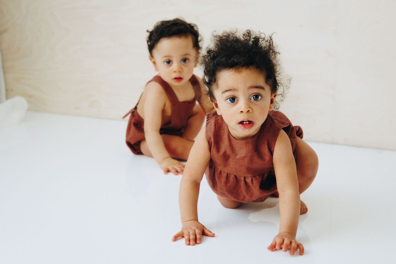 Two adorable twin toddlers in matching rust-colored rompers, one crawling and one sitting on a white floor.