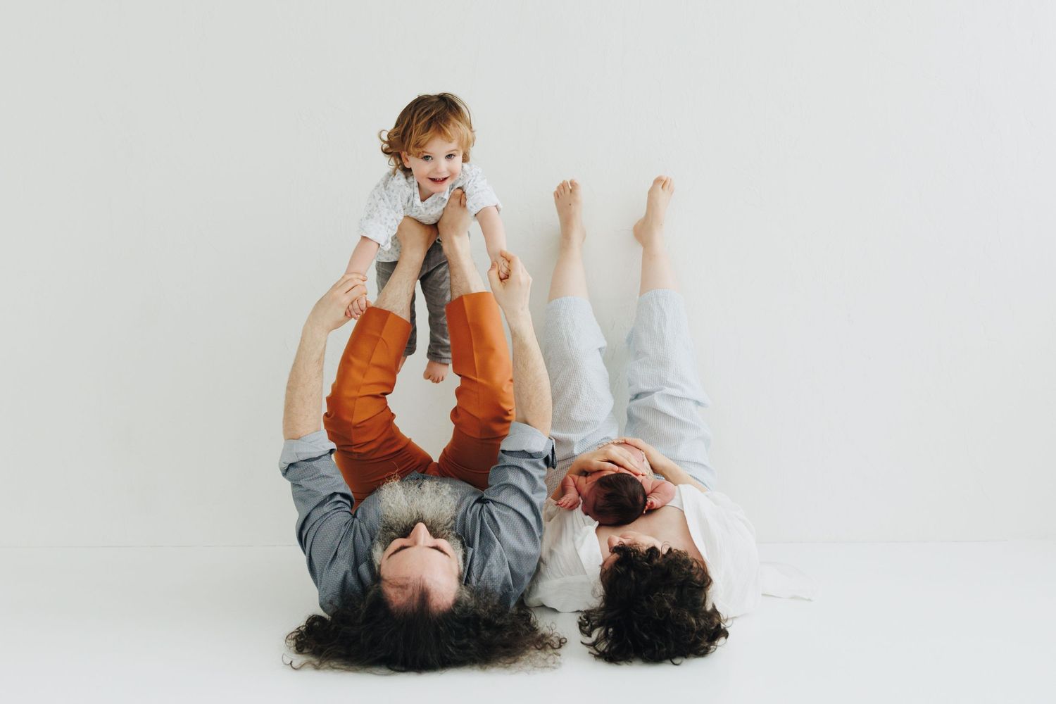 Two parents lying on floor lifting smiling baby in air in bright white studio family photo. Newborn kuvaus ja perhekuvaus Helsingissä.