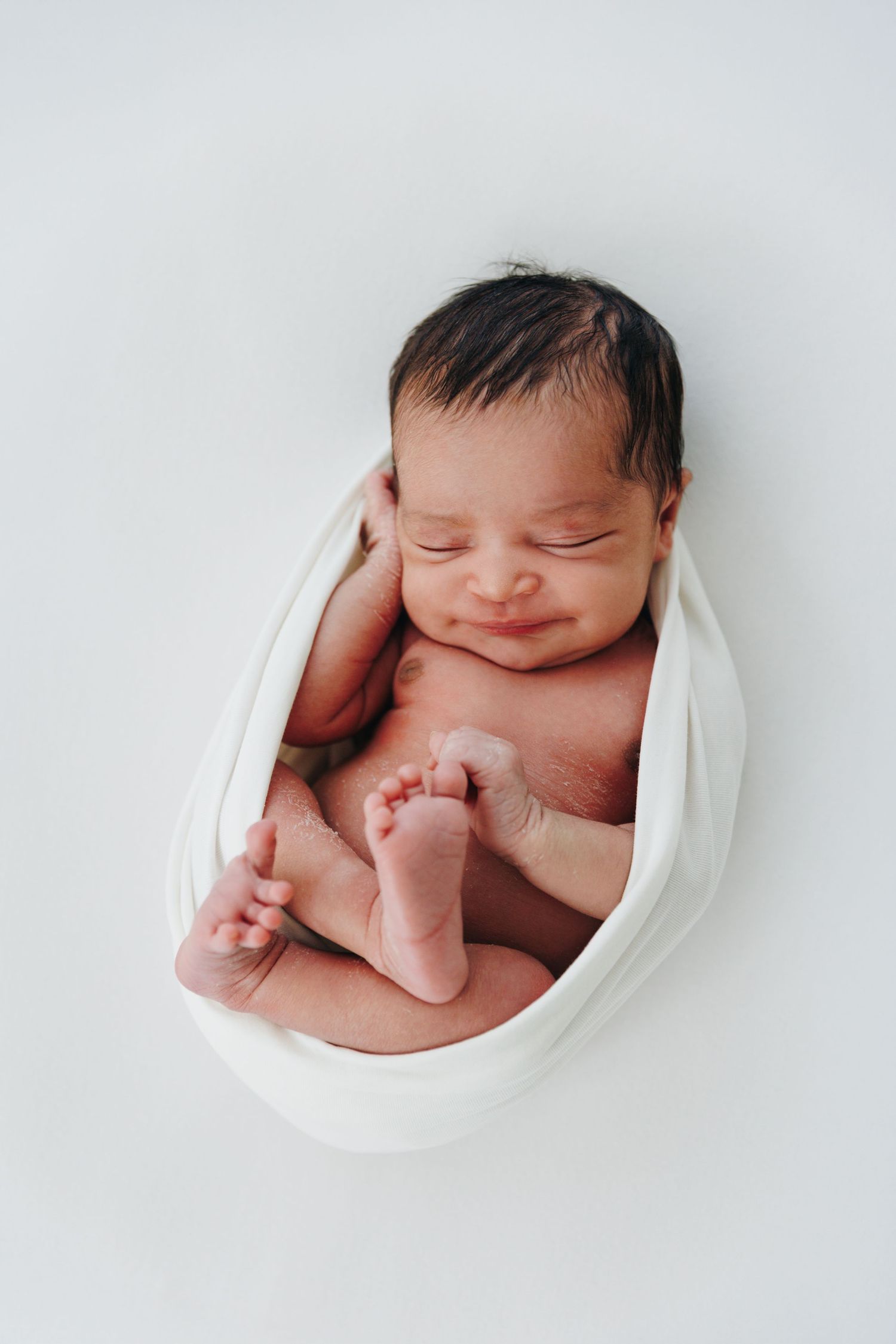 Smiling newborn baby with dark hair wrapped in white cloth, sleeping peacefully on a white background.