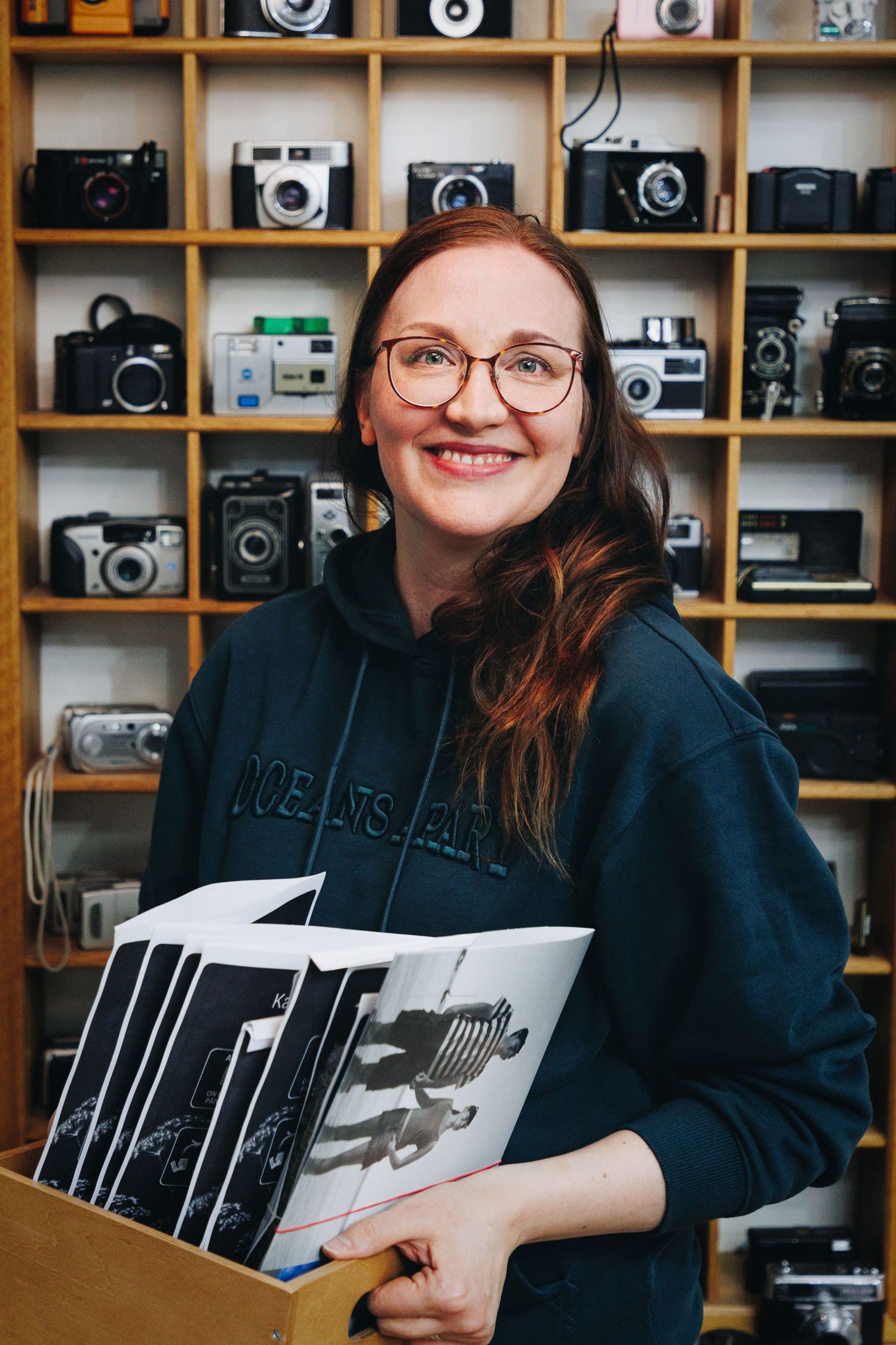 Smiling woman with glasses holds black and white photos in front of shelves displaying vintage camera collection.