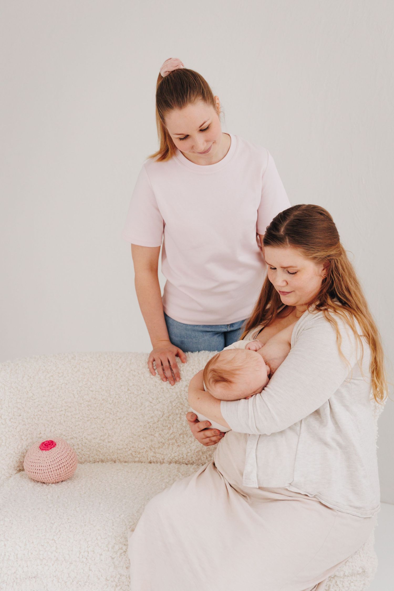 A woman breastfeeds a newborn while another woman stands beside her on a white sofa in a bright studio.