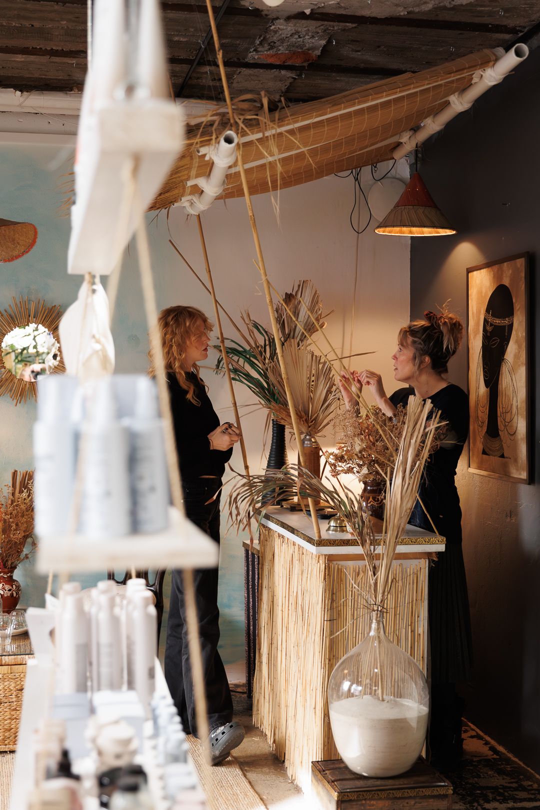 Two stylists in black outfits arranging dried palm decor at a boho-chic hair salon reception desk.