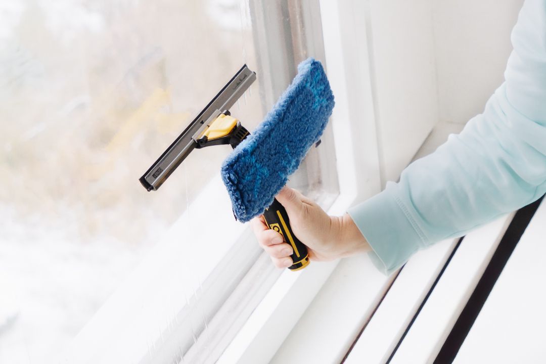 Hand holding a window squeegee and blue microfiber scrubber cleaning a white window frame indoors.