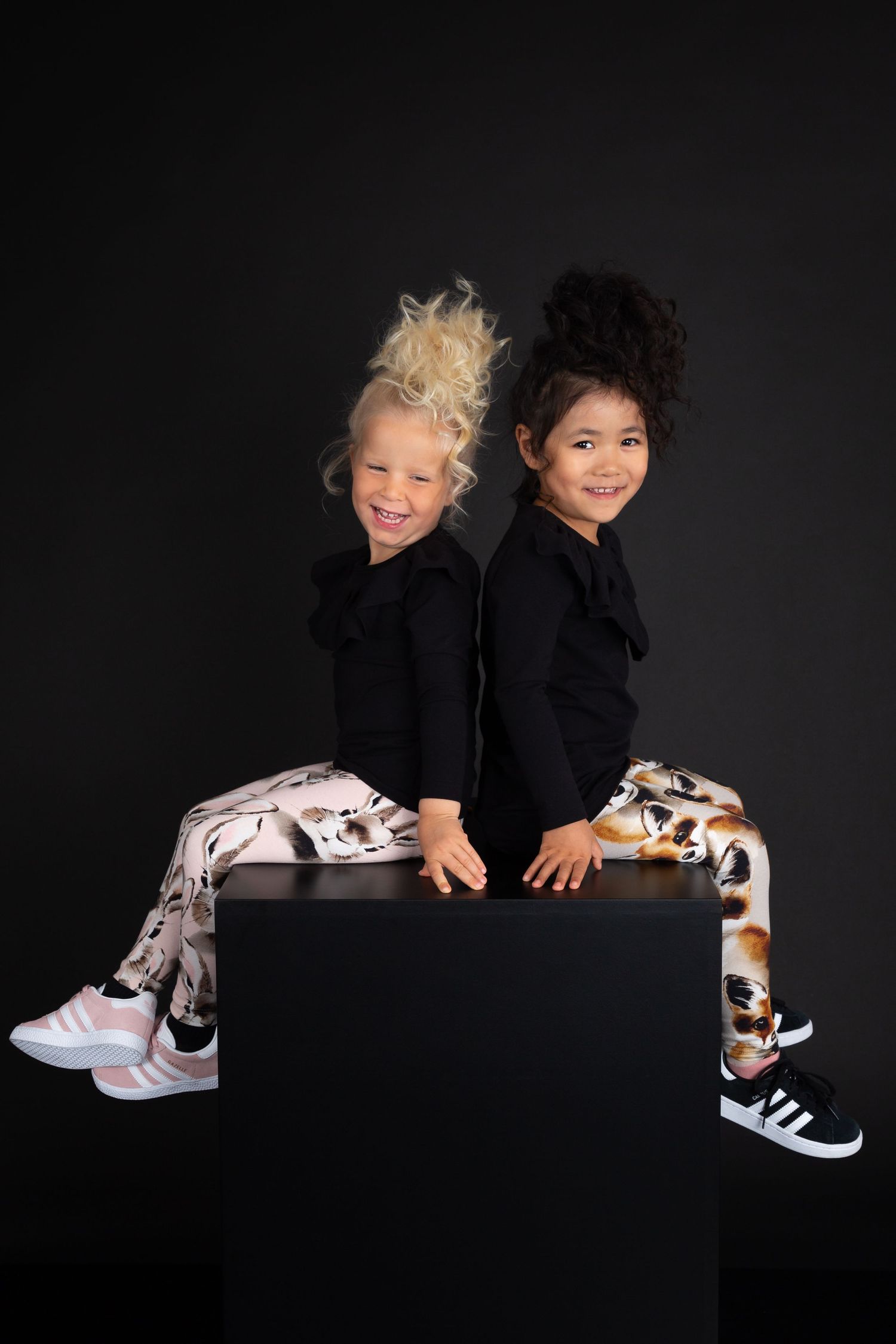 Two young girls in black tops and printed leggings sit back-to-back on a black cube against a dark background.