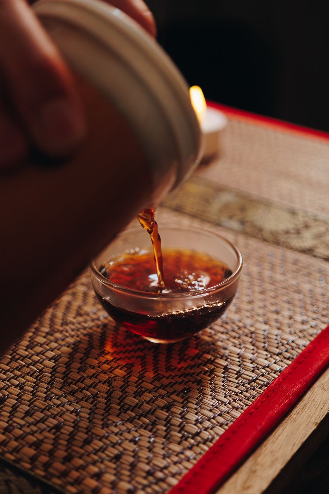 Dark amber tea being poured from ceramic gaiwan into small glass cup on woven bamboo tea tray.