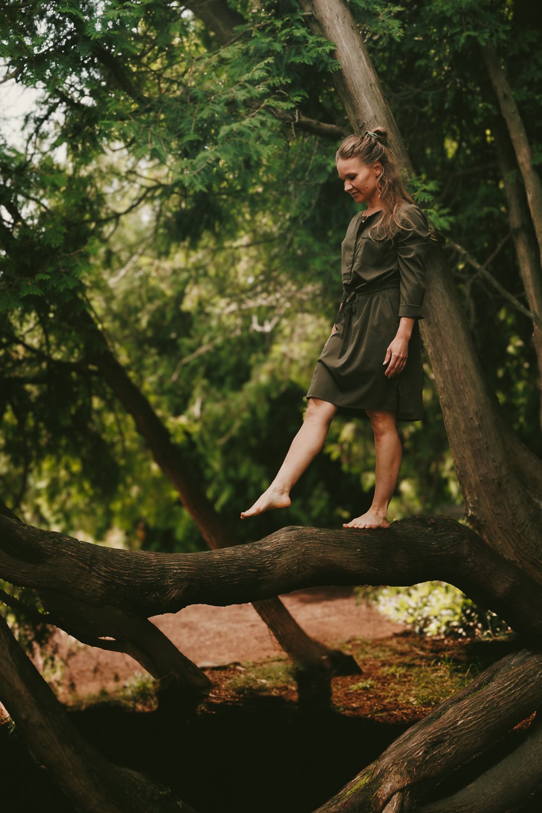 Barefoot young woman in olive dress balancing on tree branches in a lush green forest.