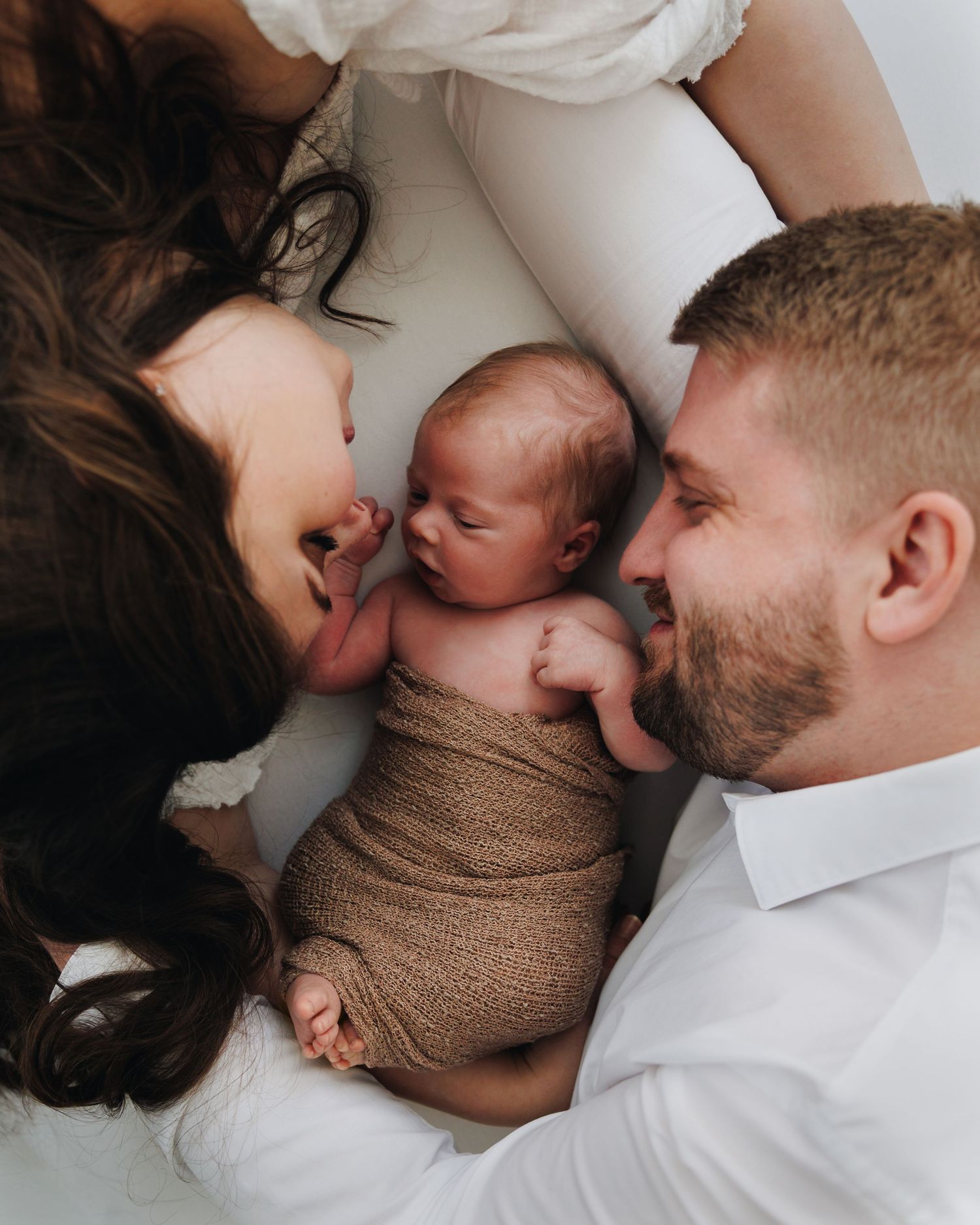 A couple lies down facing their newborn baby wrapped in a brown swaddle during a professional newborn photo session.