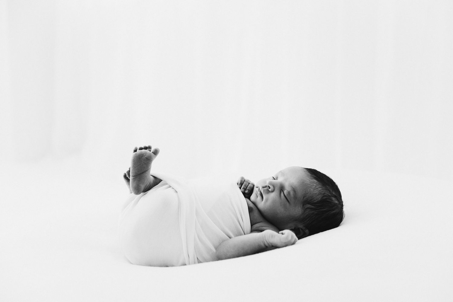 Black and white newborn photo of a swaddled baby lying on a white blanket with tiny feet raised.