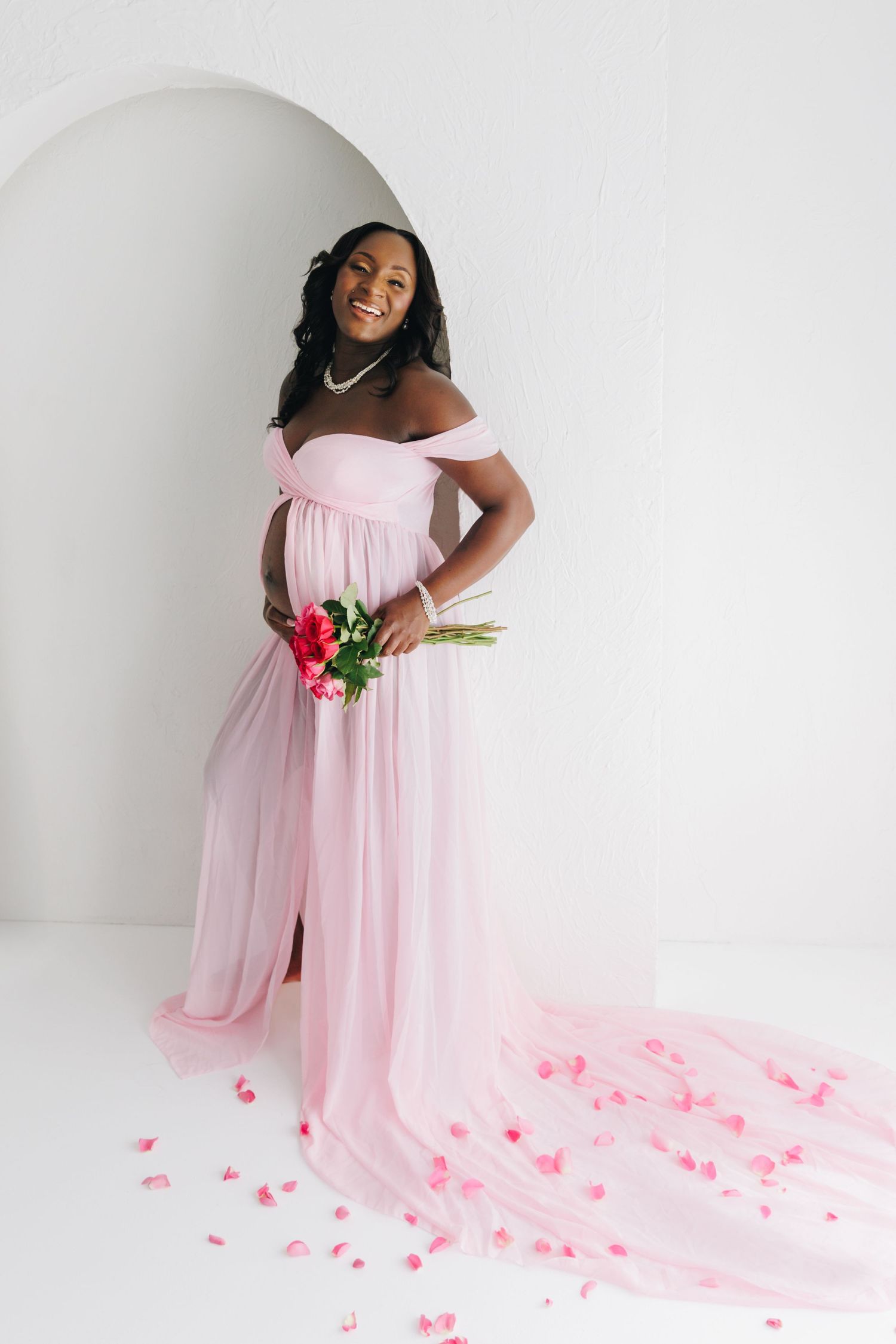 Smiling pregnant woman in pink off-shoulder tulle maternity gown holding red flowers, standing in white studio.