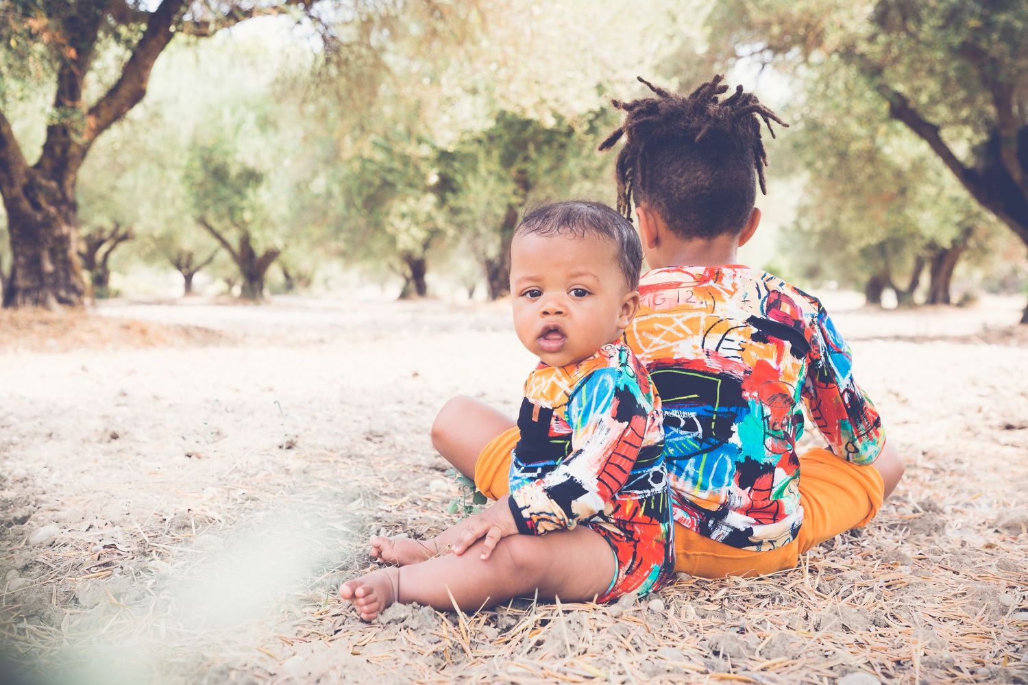 Two young children sitting together outdoors under olive trees on sandy ground, wearing colorful clothing.