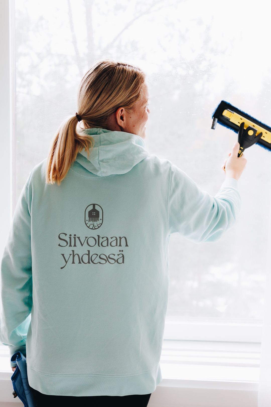 Woman in mint green Siivotan yhdessä hoodie cleaning a window with a squeegee.