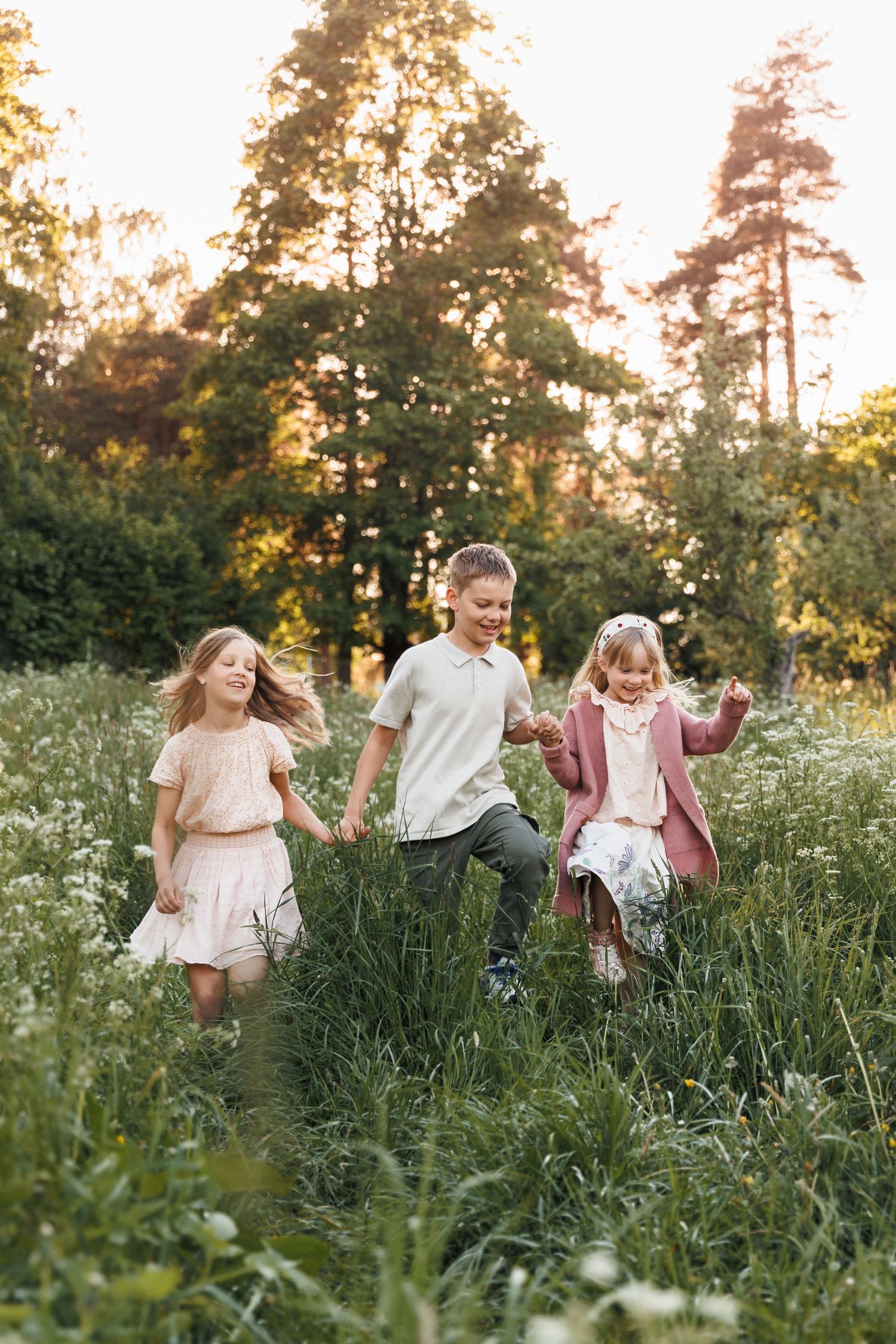 Three happy children running through a wildflower meadow at golden hour sunset with tall trees in background.