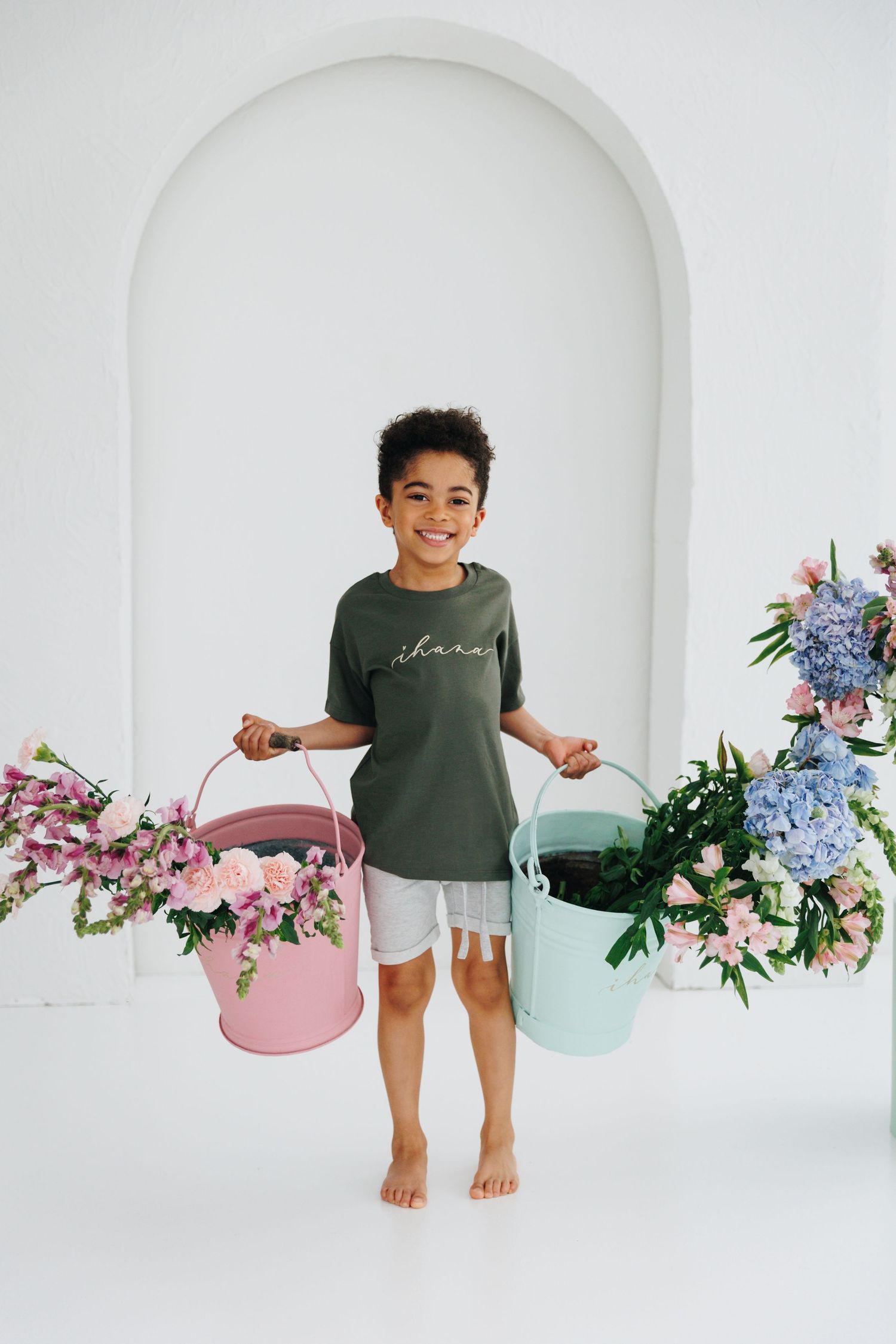 Smiling child holding pink and mint flower buckets in white studio with floral arrangements and arched backdrop.