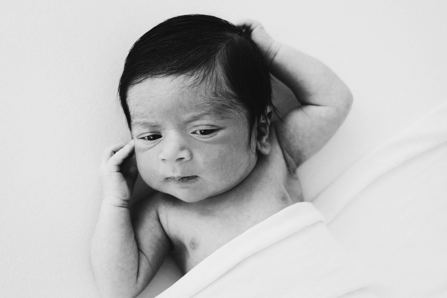 Black and white portrait of a newborn baby with dark hair, resting peacefully with hand raised near face.