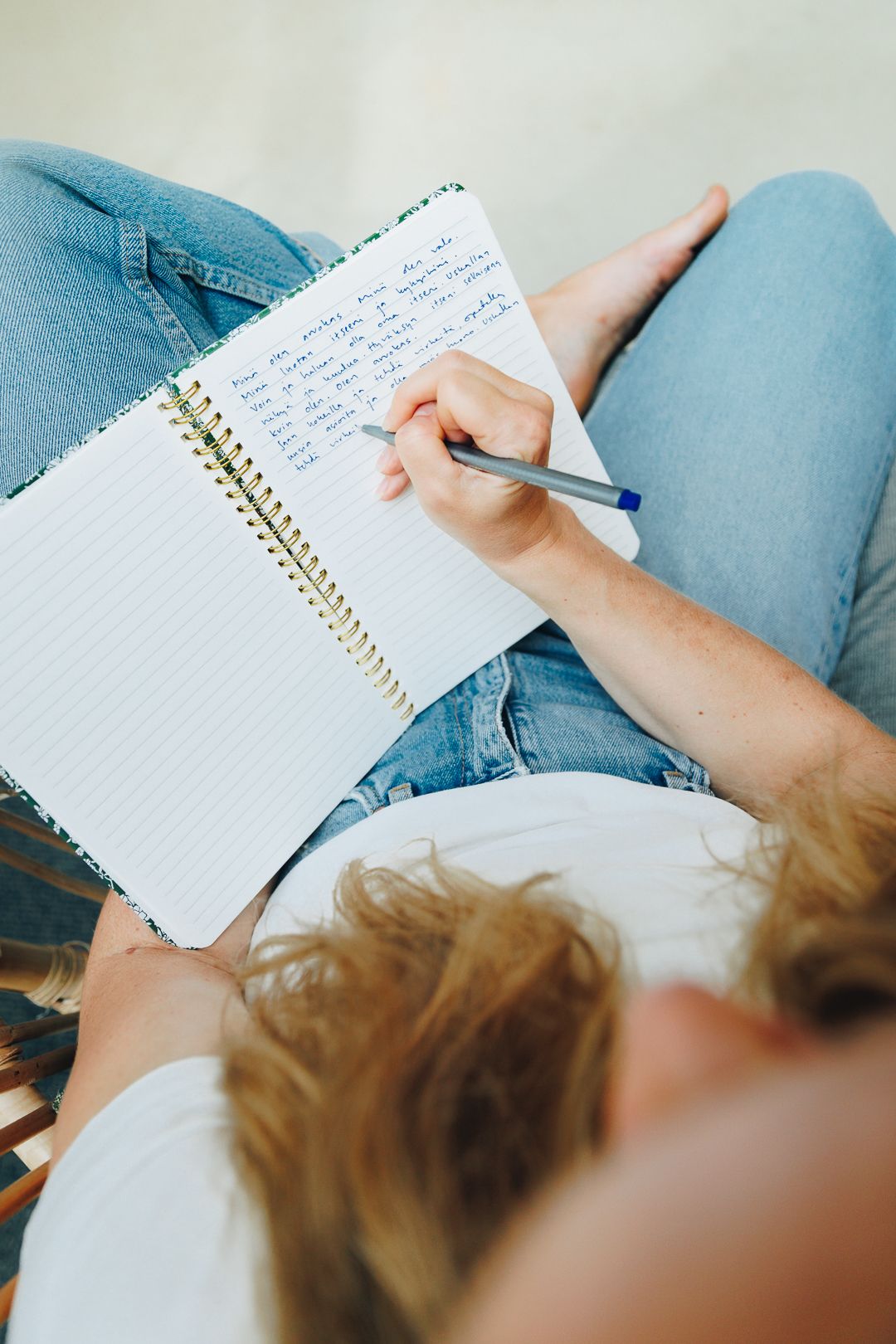 Person writing notes in a spiral notebook while sitting cross-legged, wearing blue jeans, holding a pen.