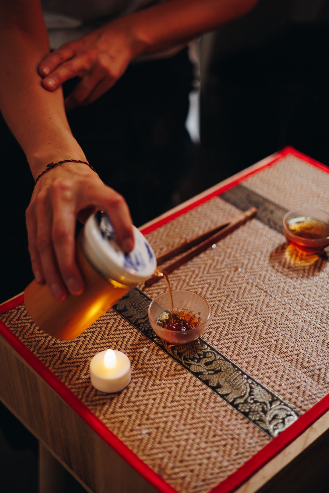 Person pouring tea into a small glass cup during a traditional Chinese tea ceremony with candles.