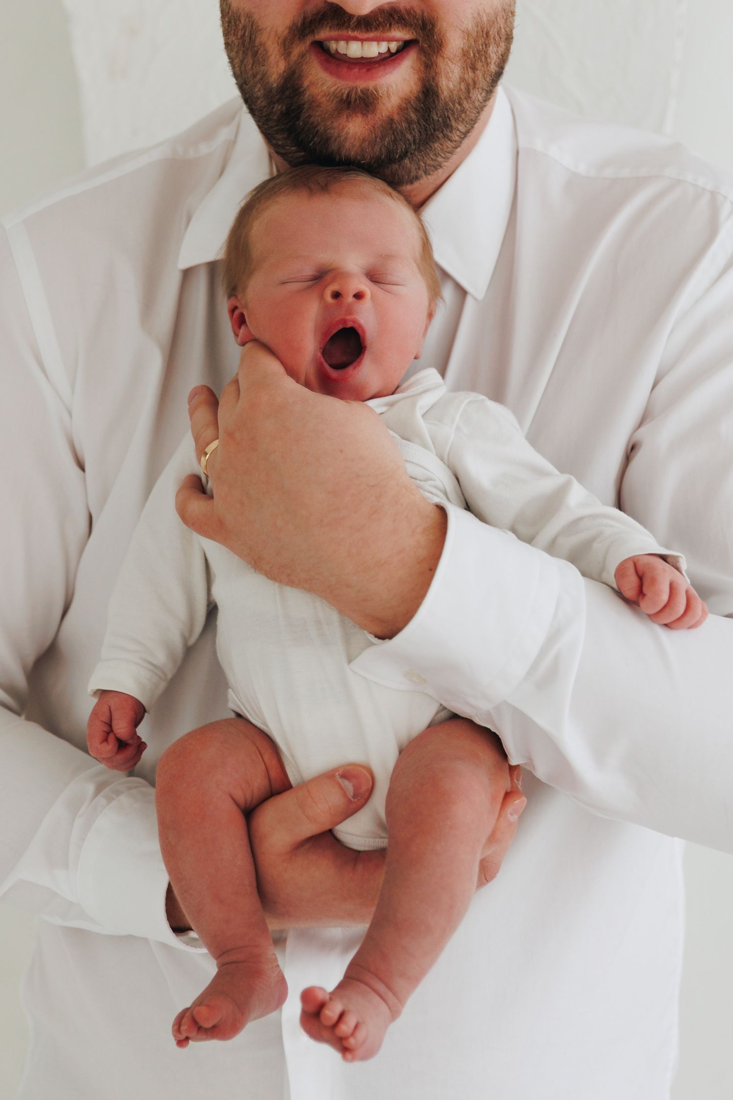 Smiling bearded father in white shirt holding a yawning newborn baby dressed in white.