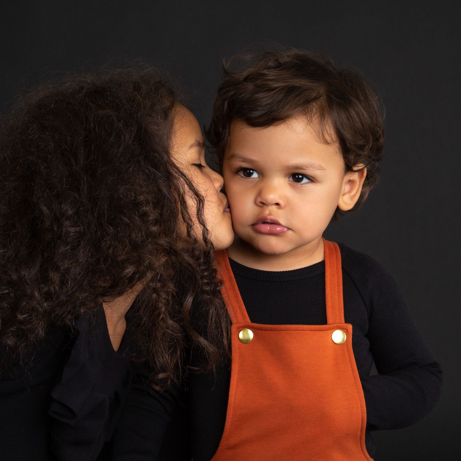 A young toddler in an orange overall poses and plays while being photographed in a dark studio setting.