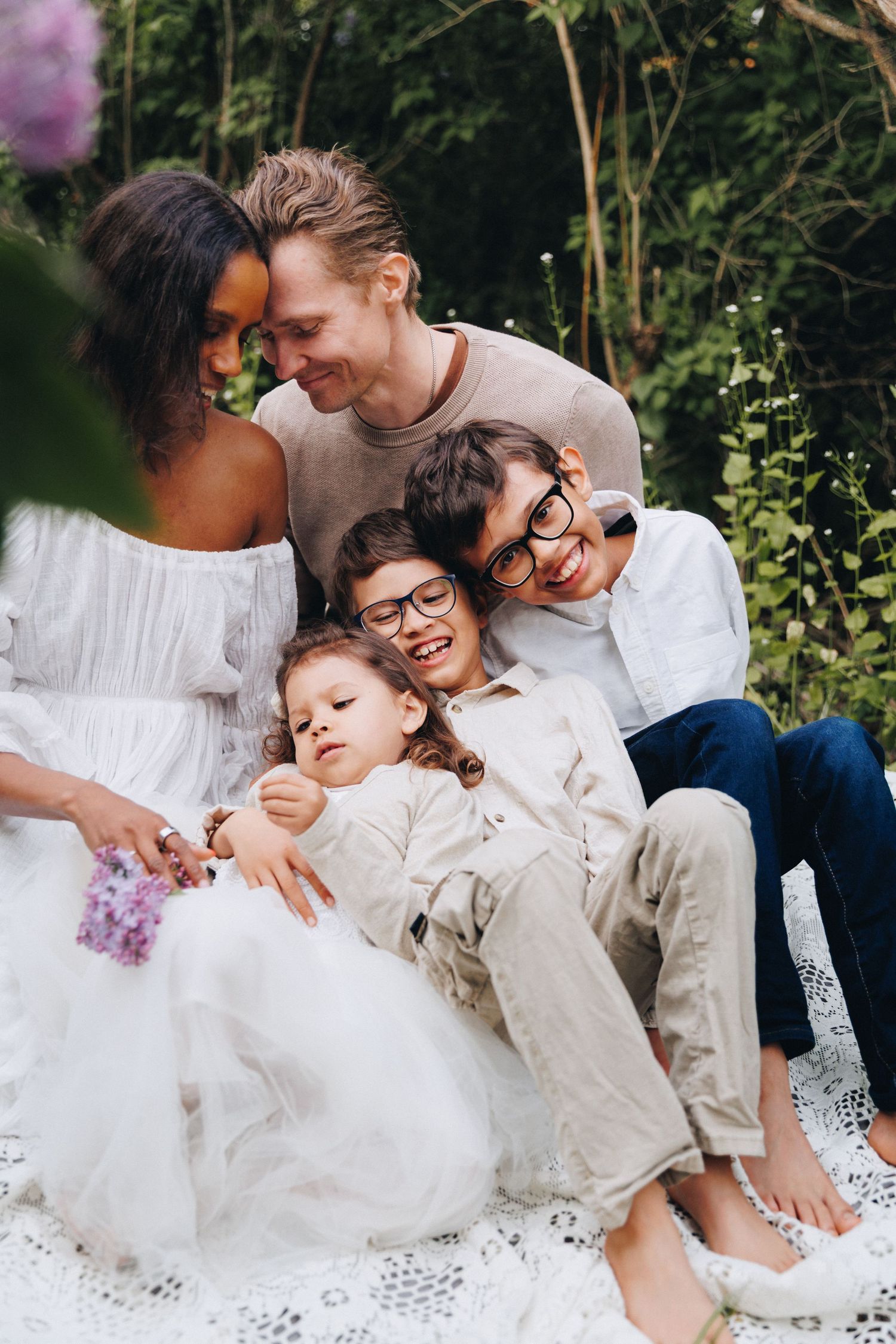Happy family with two parents and three children sitting together outdoors on a blanket surrounded by greenery.