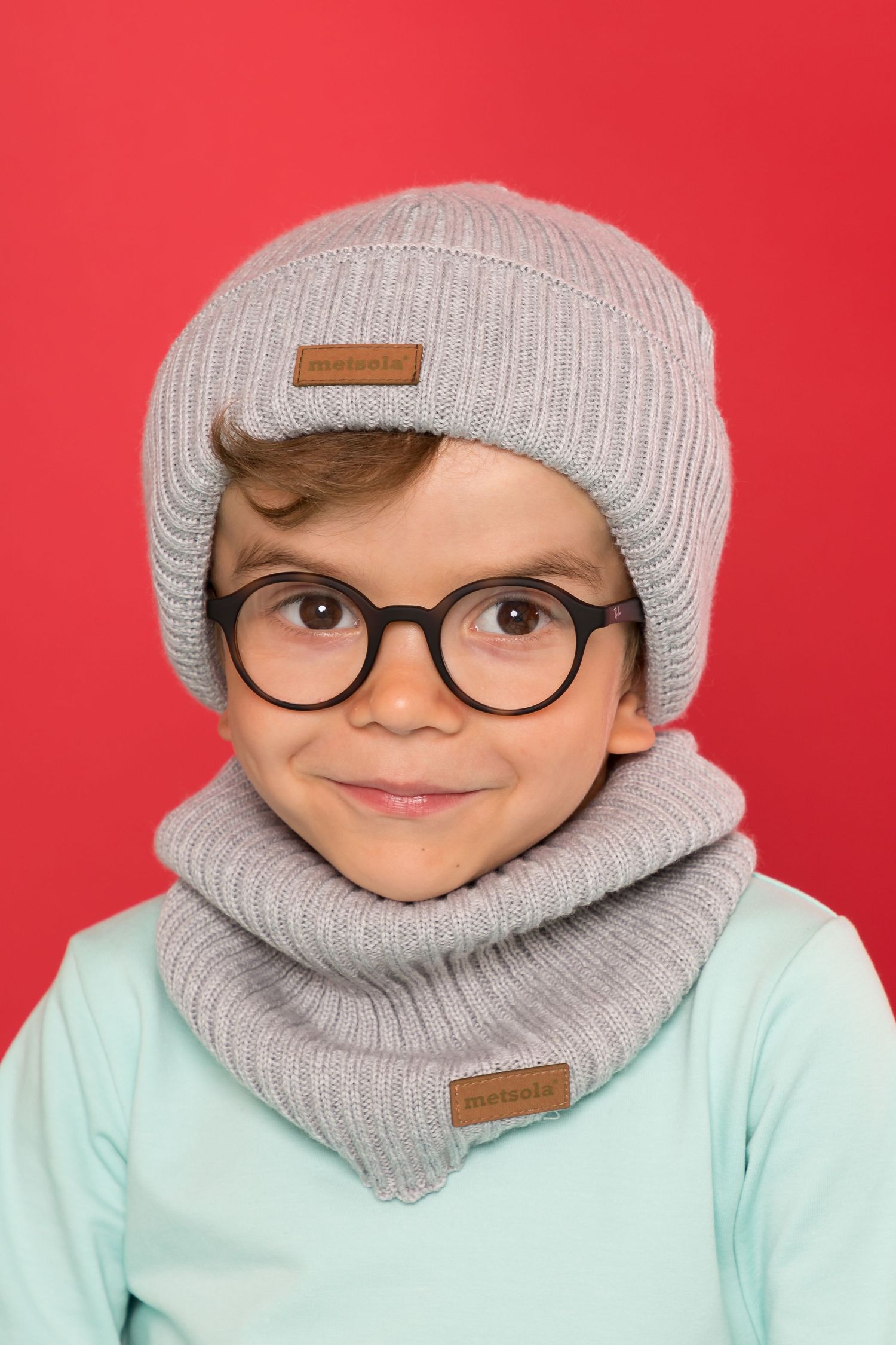 Cute child wearing gray knit beanie hat, scarf, and round glasses, smiling against a red background.