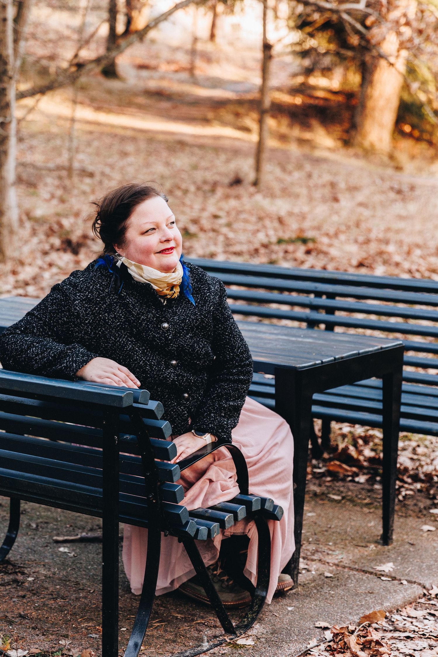 Smiling middle-aged woman in black jacket and pink skirt sitting on a park bench surrounded by autumn leaves.
