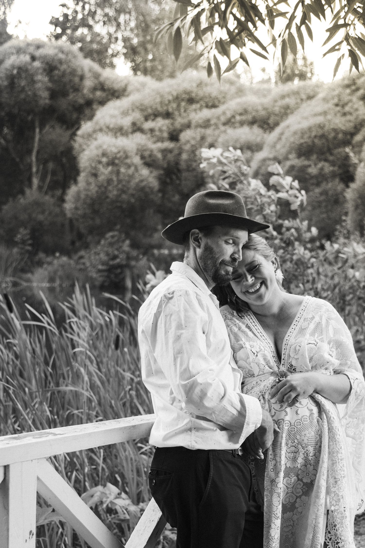 Black and white photo of a laughing couple embracing on a bridge in a lush garden, man wearing a hat.