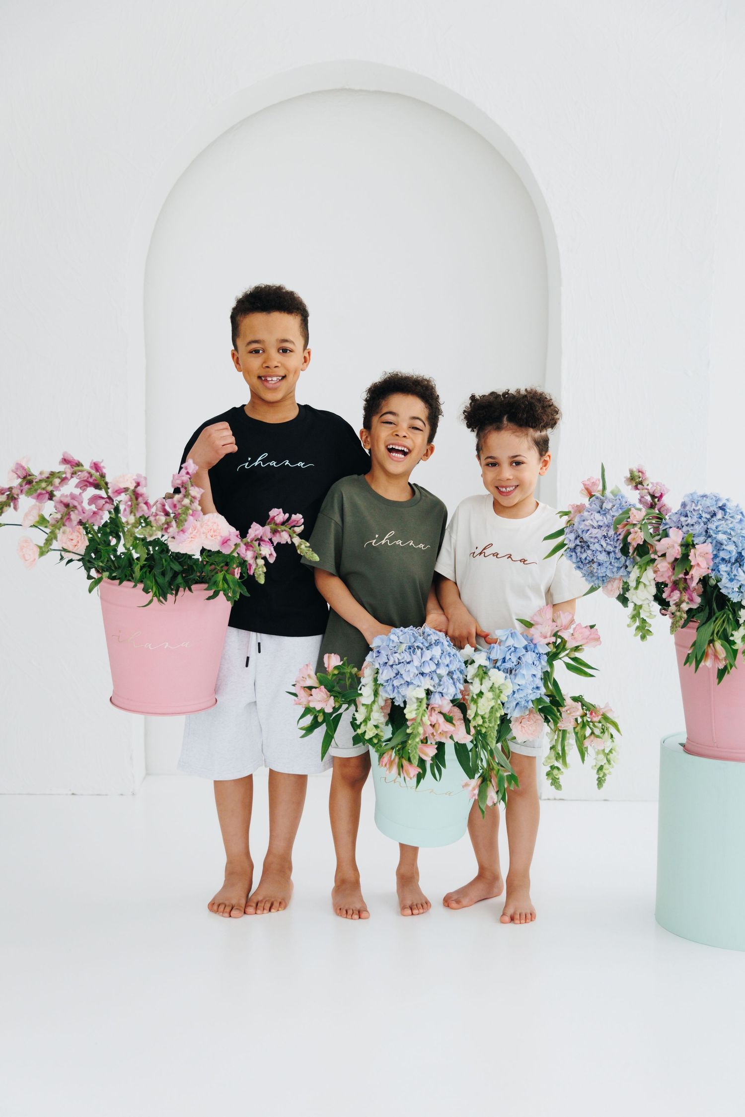 Three happy kids holding colorful flower bouquets in pastel buckets, wearing matching shirts in a bright white studio.
