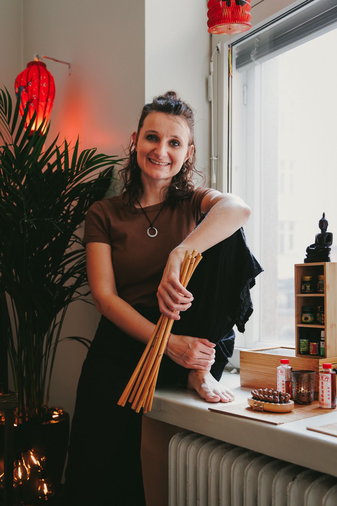 Smiling female massage therapist holding bamboo sticks in a warmly decorated Asian-inspired studio.