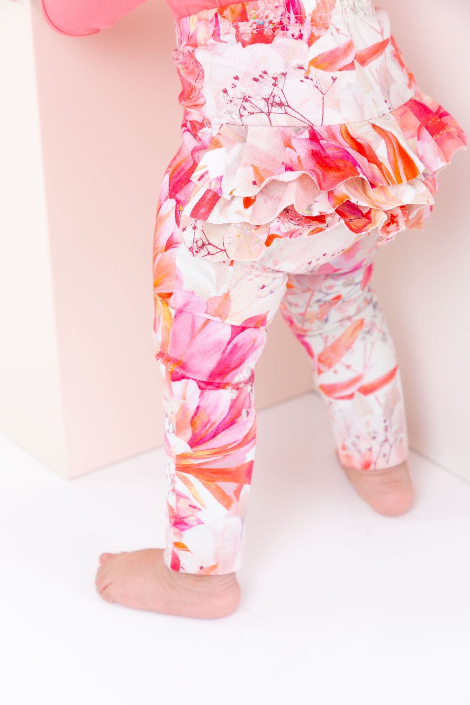 Baby girl wearing pink floral ruffle leggings and matching top, standing barefoot against a white background.