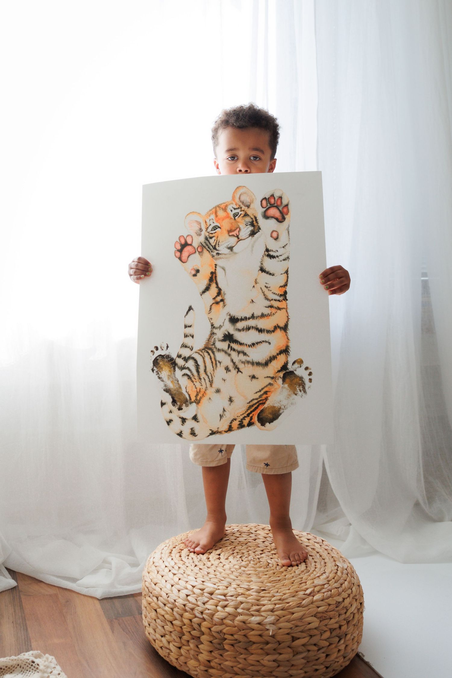 A young child stands on a wicker pouf holding a large tiger illustration print in a bright white room.