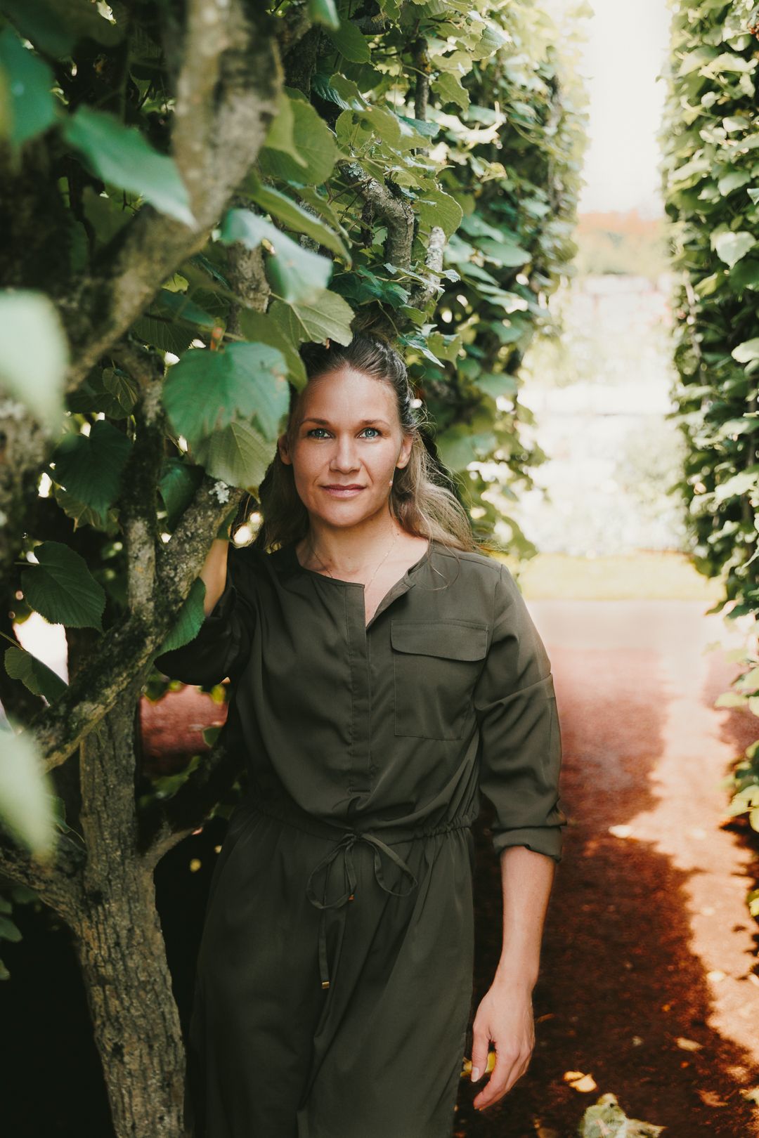 Woman in olive green jumpsuit standing among lush green trees in a sunlit garden pathway.
