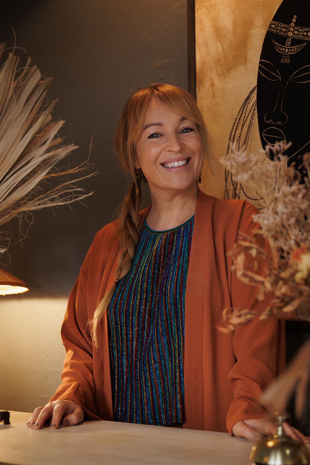 Smiling woman with red hair in orange blazer stands at counter in warmly decorated salon interior.