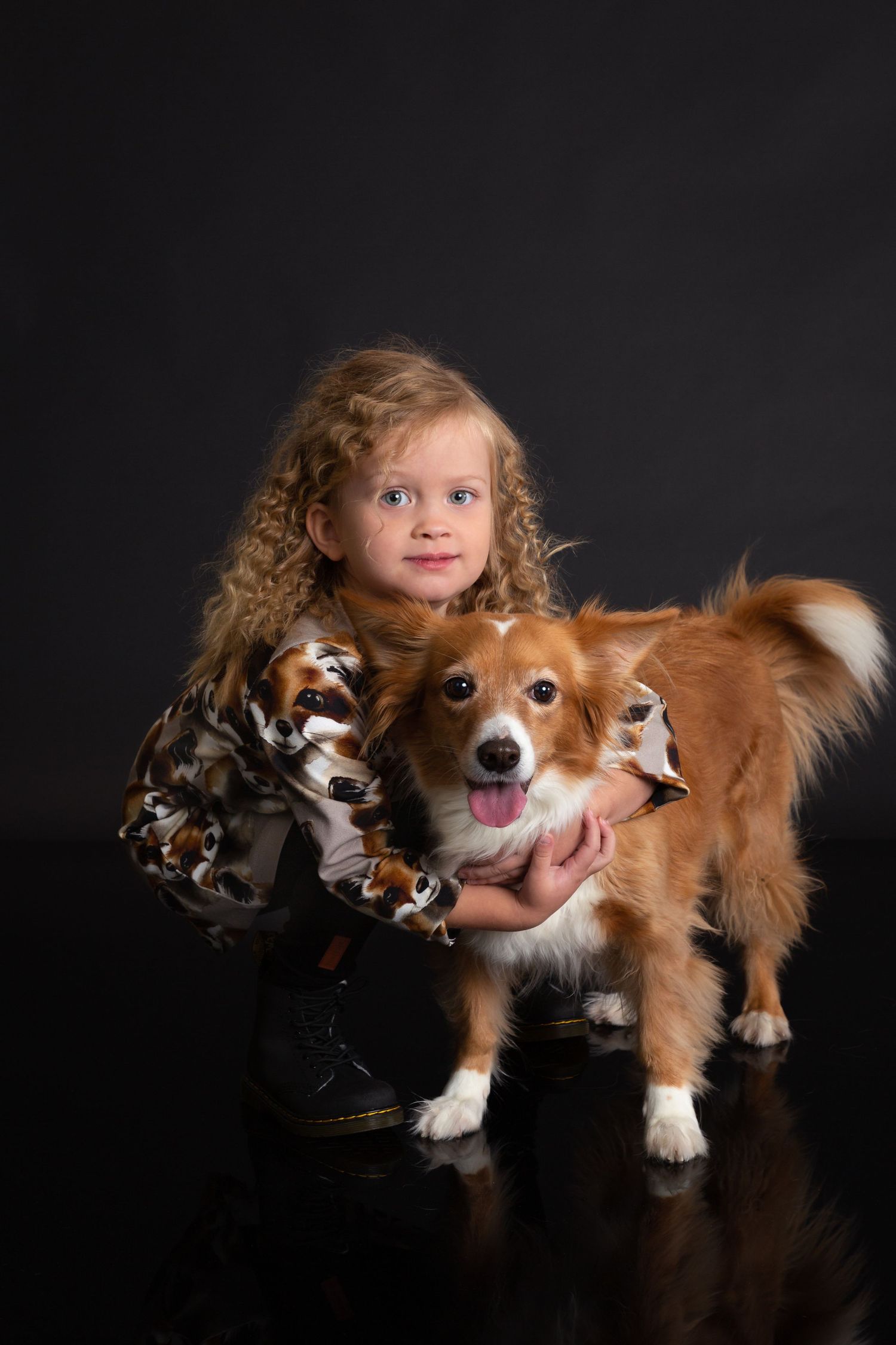 A curly-haired girl in a printed jacket hugs a happy Corgi dog against a dark studio background.