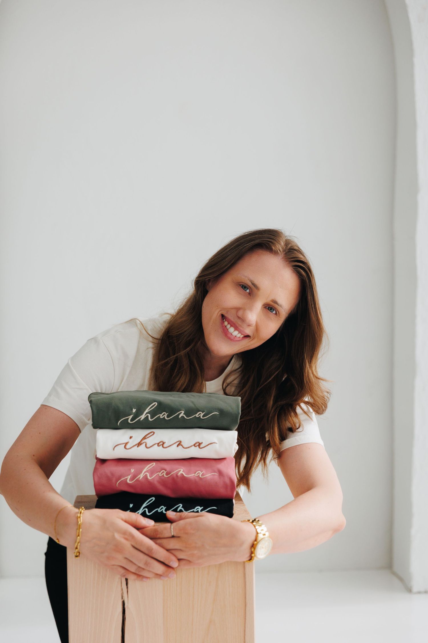 Smiling woman holding a stack of folded ihara branded sweatshirts in green, white, and burgundy colors.