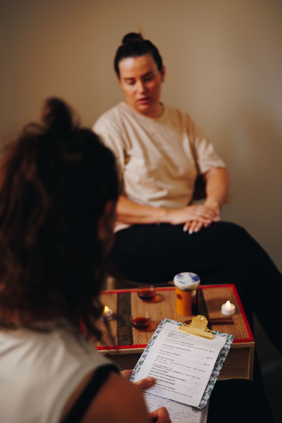 Woman holding menu clipboard during traditional tea ceremony consultation with practitioner seated opposite.