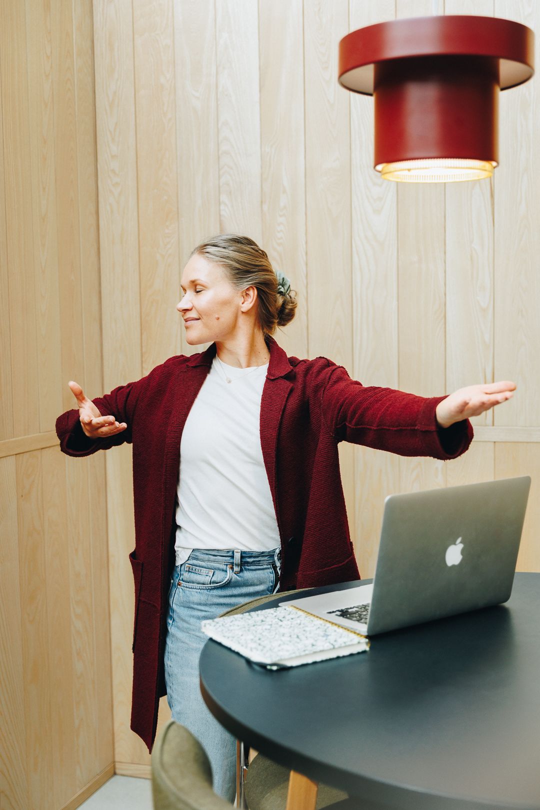 Woman in red blazer spreads arms wide, smiling at desk with laptop, beside a red pendant lamp.