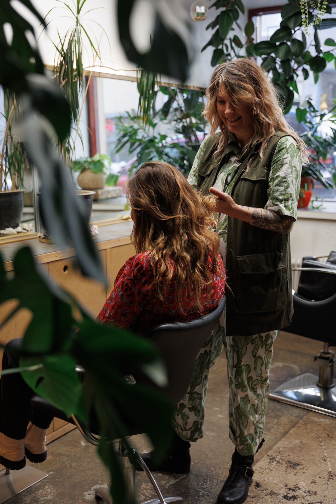 A smiling hairstylist in a green outfit works on a client's wavy hair in a lush, plant-filled salon.