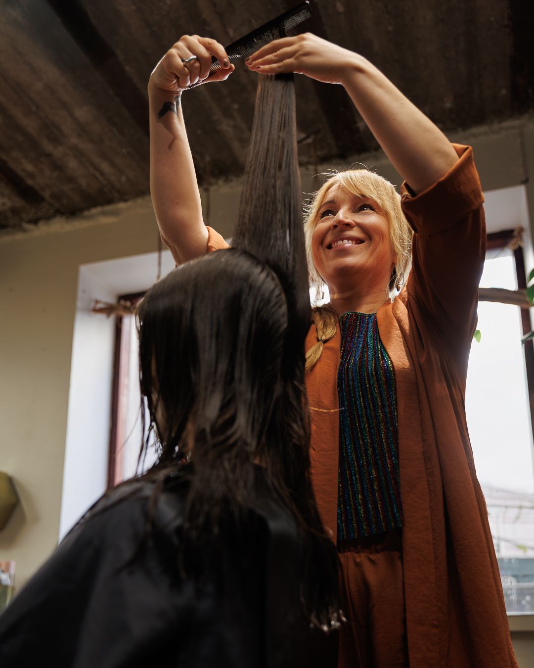 A smiling blonde hairstylist cuts a client's long dark hair with scissors raised above her head.