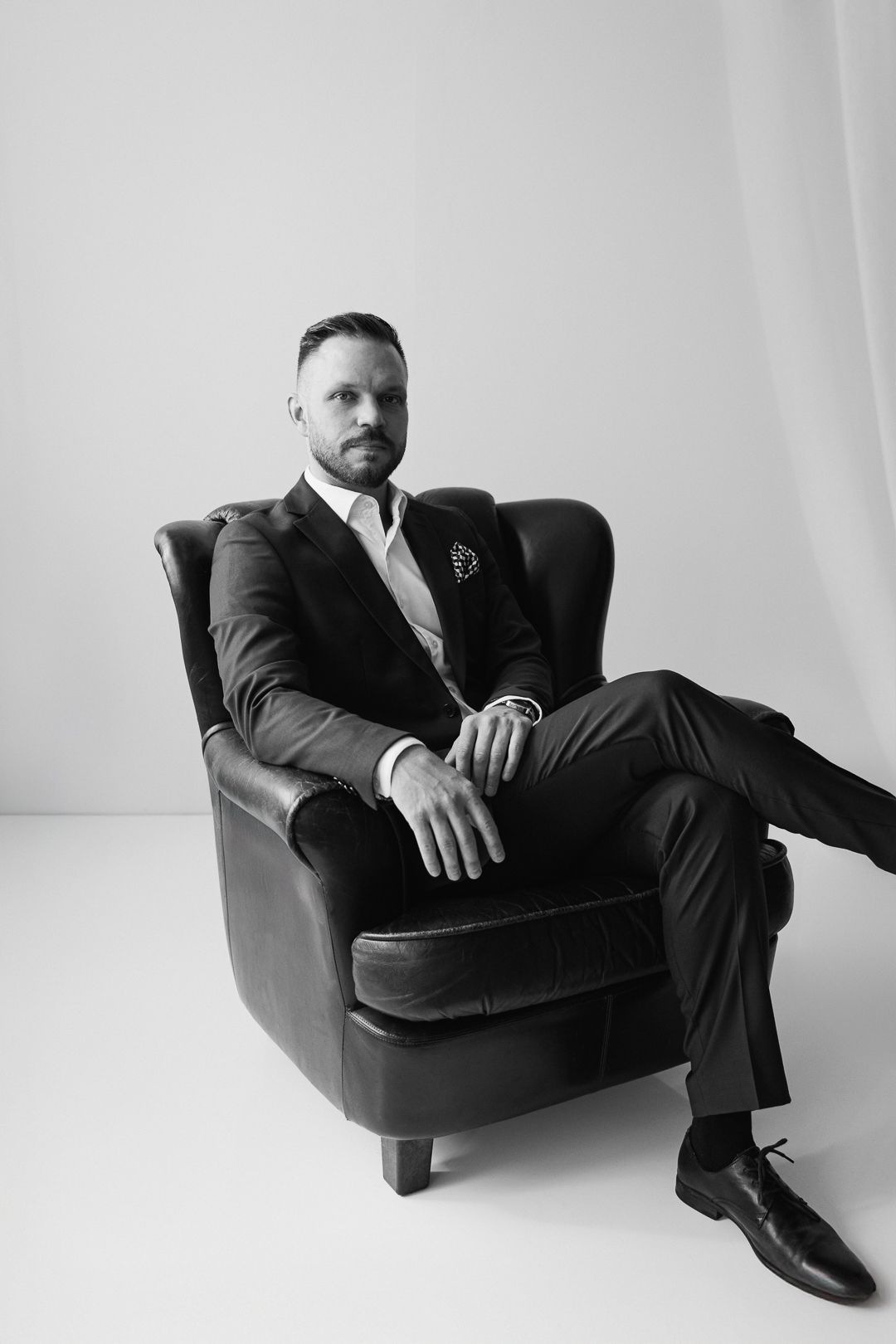 Black and white portrait of someone in a suit sitting elegantly in a dark leather armchair against white background.