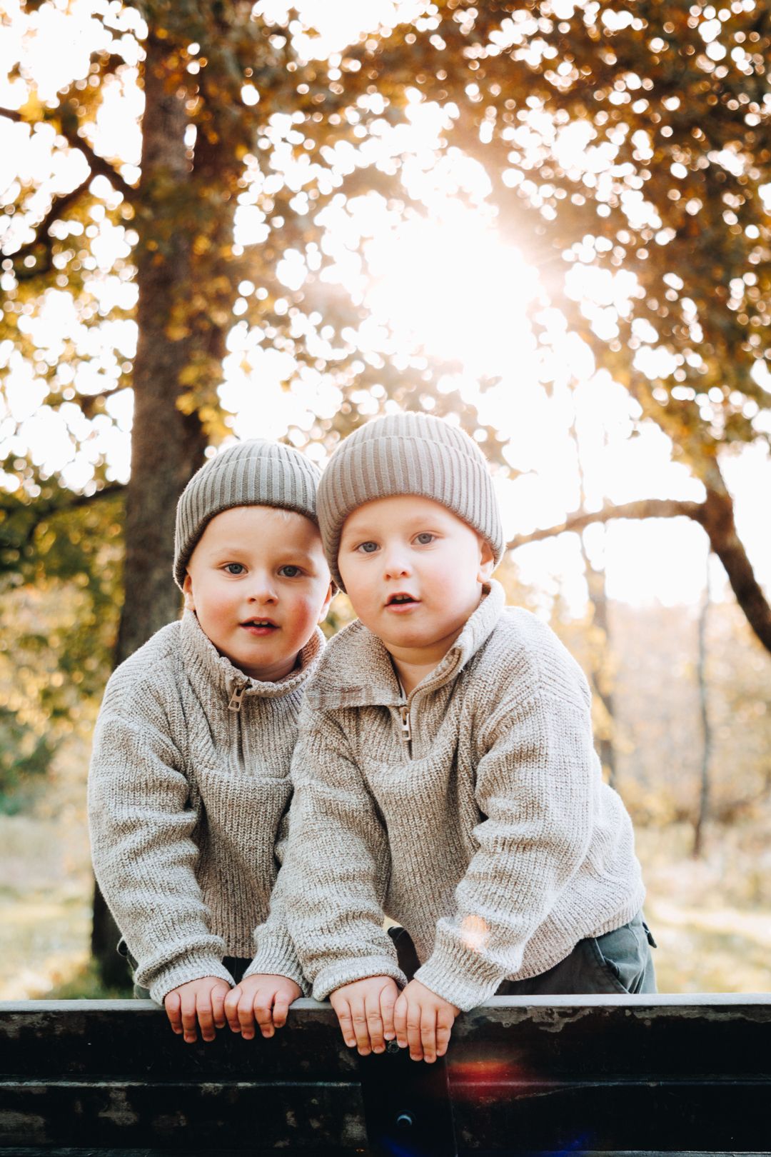 Two children in matching gray sweaters and beanies lean together against a railing during golden hour sunset.