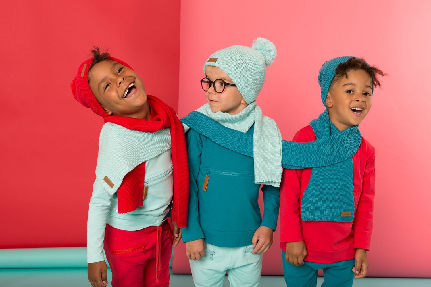 Three cheerful kids wearing colorful winter outfits with scarves and hats pose against a bright pink and teal background.