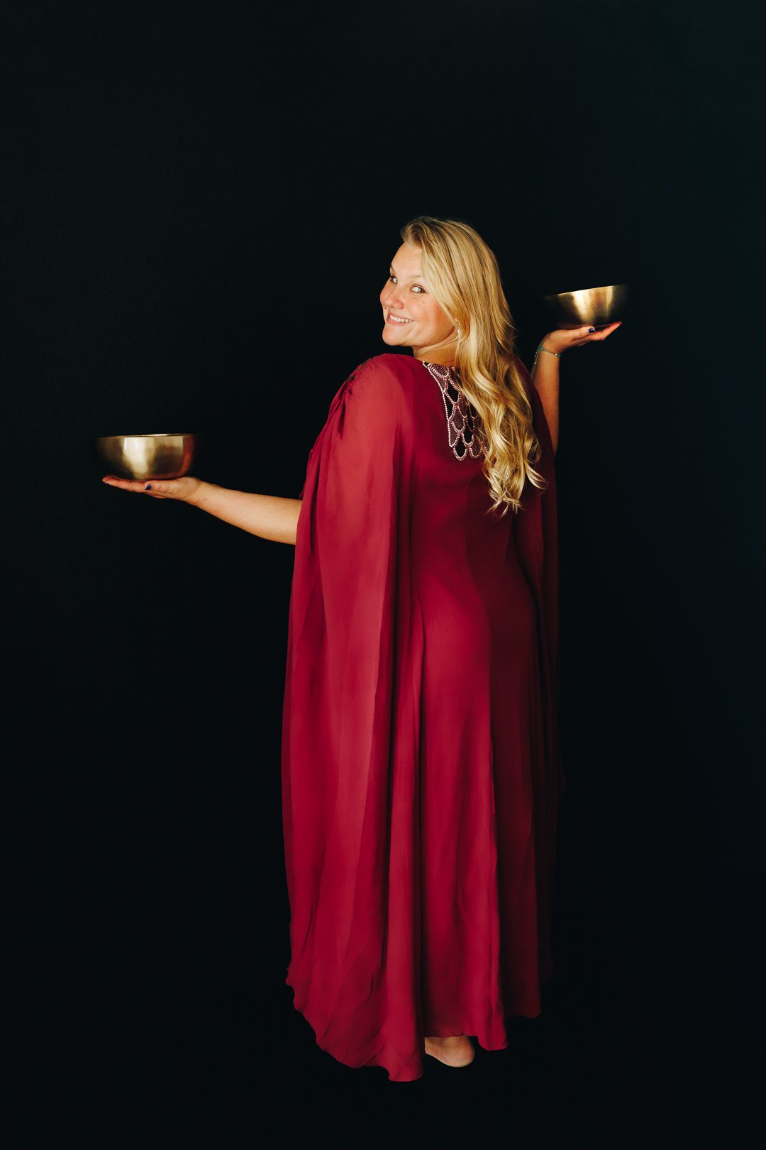 Smiling blonde woman in red cape gown holding two golden singing bowls against black background.
