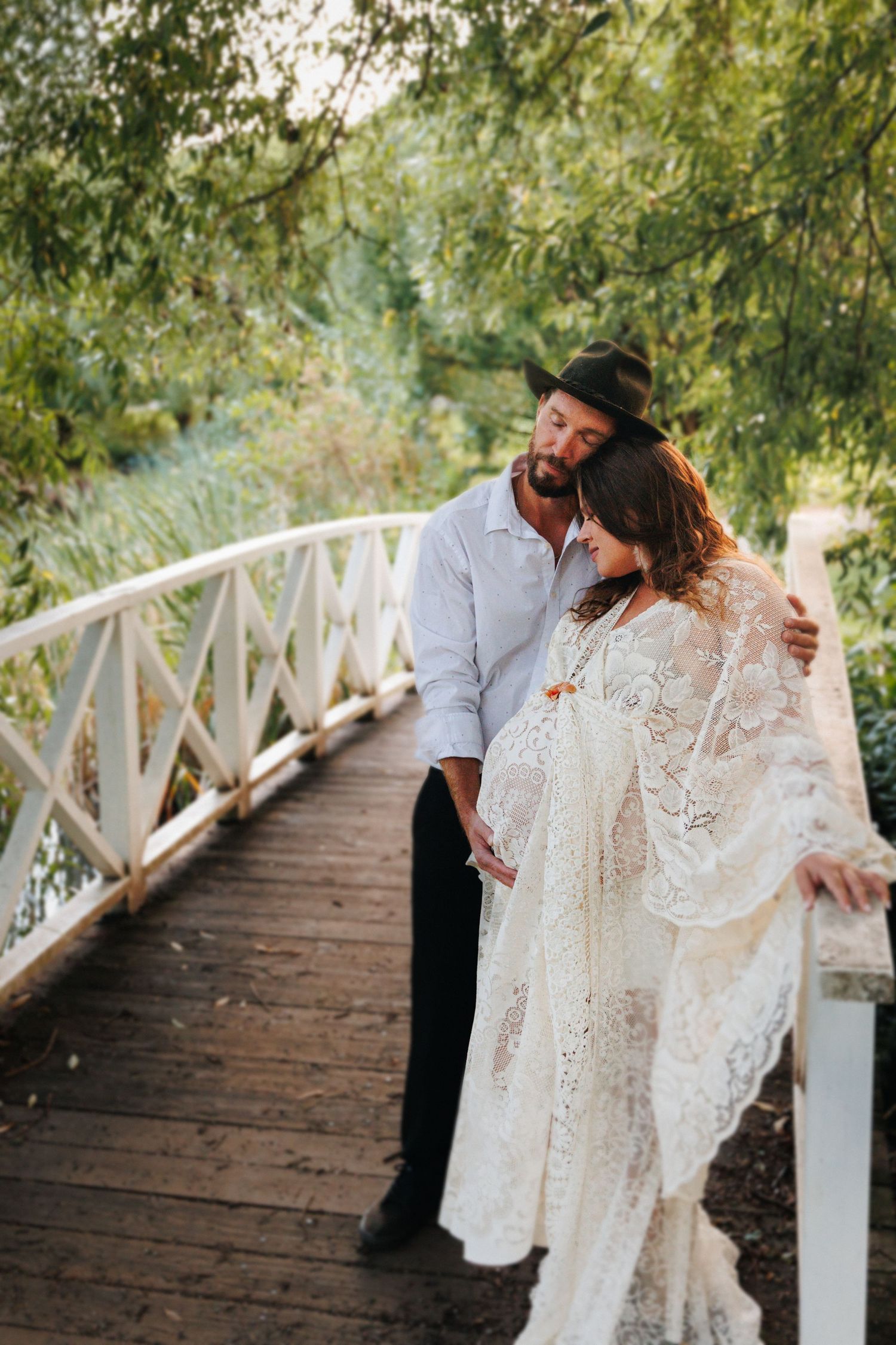Couple on a rustic wooden bridge, man in hat embraces pregnant woman in white lace dress, surrounded by lush greenery.