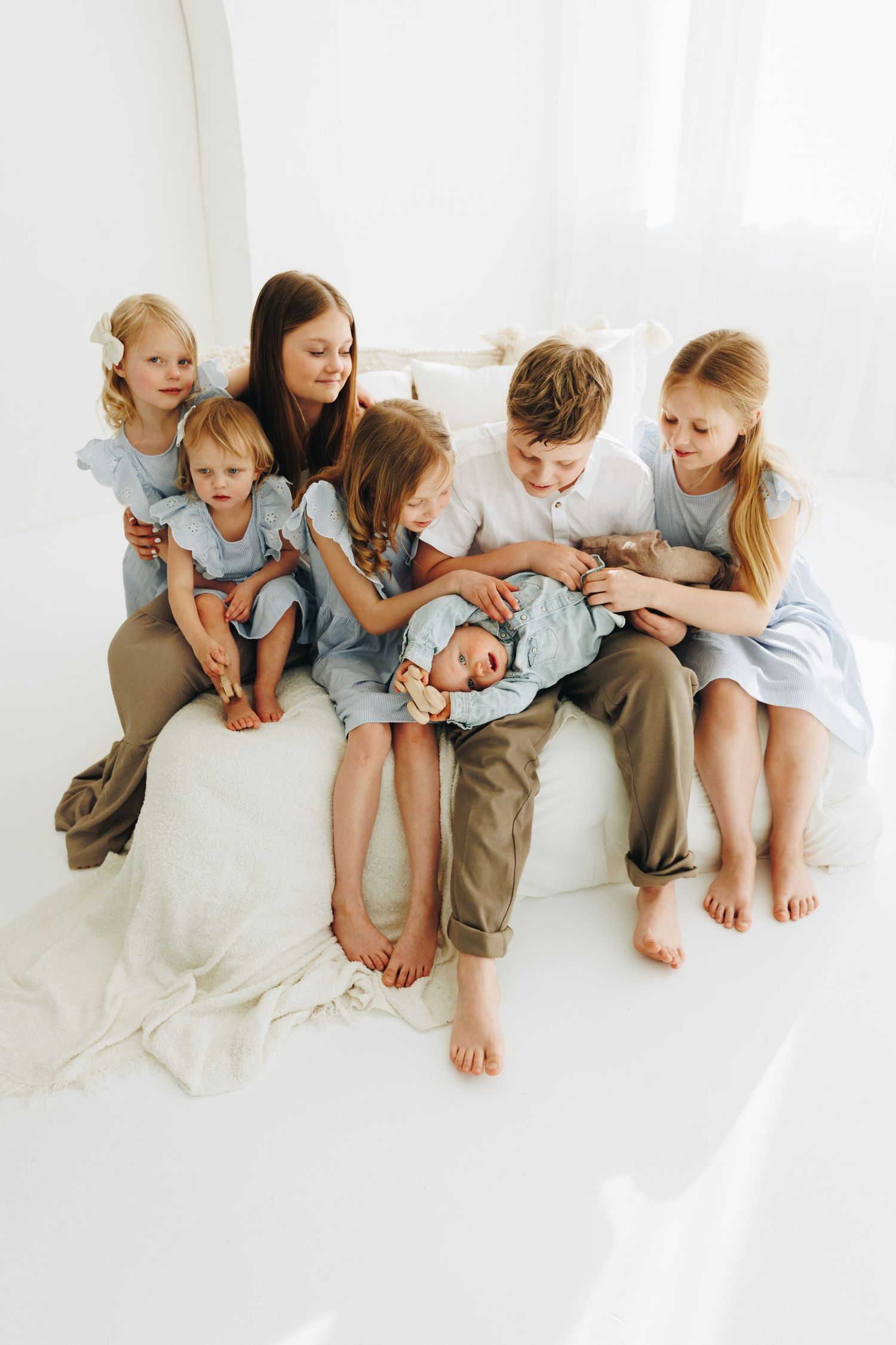 A mother sits on a white bed surrounded by her six children, including a newborn baby, in a bright white room.