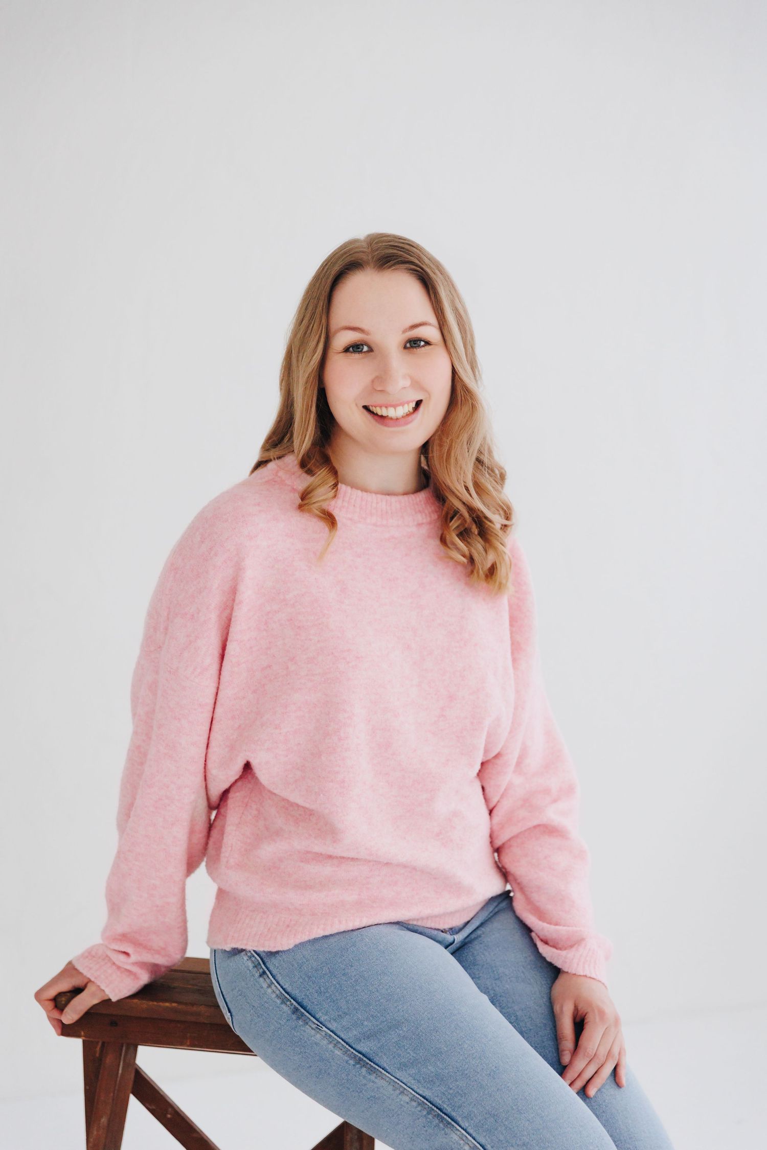 Smiling woman in a pink sweater and jeans sitting on a wooden stool against a white background.