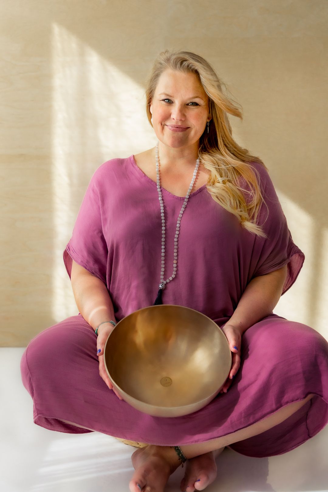 Woman in purple outfit sitting cross-legged holding a golden singing bowl with a mala bead necklace.